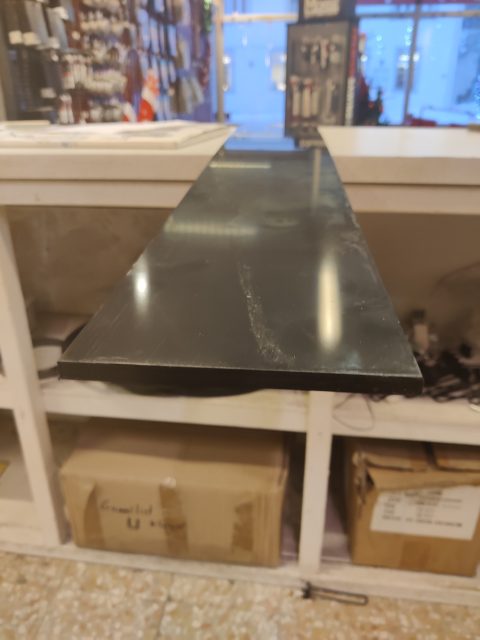 En närbild av en Skidplast 2000 x 163 x 10 mm svart 1:a bänkskiva med synliga ränder och reflektioner, med utsikt över förvaringshyllor med kartonger under.