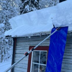 Snötäckt tak med istappar och skidstavar stödda mot en röd stuga, omgiven av snötäckta träd. Taksnöhyvel 5,85m med ovanligt kraftig oval alu.stång