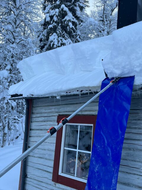 Snötäckt tak med istappar och skidstavar stödda mot en röd stuga, omgiven av snötäckta träd. Taksnöhyvel 5,85m med ovanligt kraftig oval alu.stång