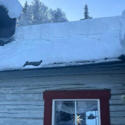 En Taksnöhyvel 5,85m med ovanligt kraftig oval alu.stång med en röd dörr delvis skymd av ett tjockt lager snö på taket och omgivande landskap.