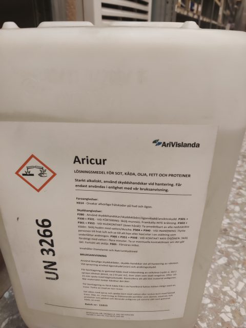Plastbehållare med 'AriCure 25Liters dunk' rengöringsmedel med varnings- och informationsetiketter, förvarad inomhus.